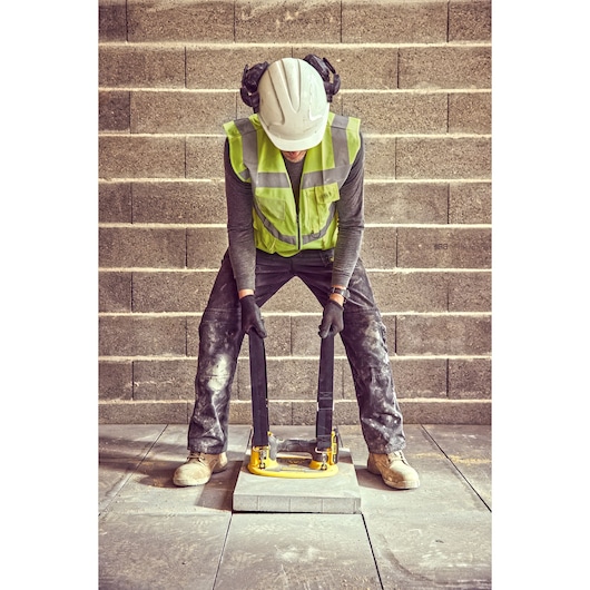 A construction worker wearing a safety helmet, ear protection, high-visibility vest, gloves, and work boots uses a yellow DEWALT lifting device to move a concrete slab, standing on a tiled surface with a brick wall in the background.