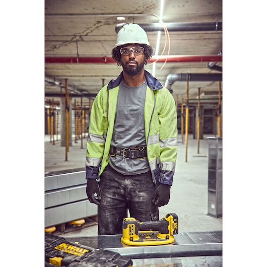 A construction worker wearing a reflective jacket, safety gloves, and a white hard hat stands in an unfinished building site. On the table in front there is a DEWALT DCE590 cordless tool and a DEWALT toolbox. The background shows pillars and ductwork in a concrete building under construction.
