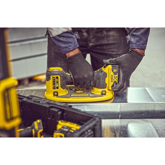 A person wearing black gloves is using a DEWALT DCE590 cordless power tool with dual suction cups to handle a sheet of metal in an industrial workspace.