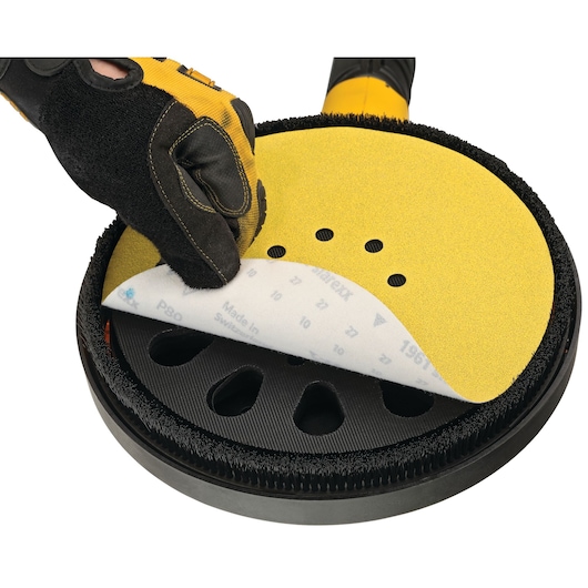 A close-up of a hand wearing a protective glove attaching a yellow sanding disc to the head of a DEWALT drywall sander.