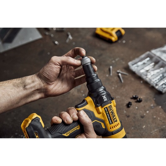 A person is holding and adjusting a DEWALT cordless power tool with a yellow and black design on a workbench, surrounded by screws and small parts.