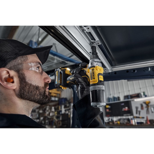A person wearing ear protection and gloves uses a DEWALT DCF414N cordless power tool to fasten or unfasten a metal component indoors, in an industrial or workshop setting.