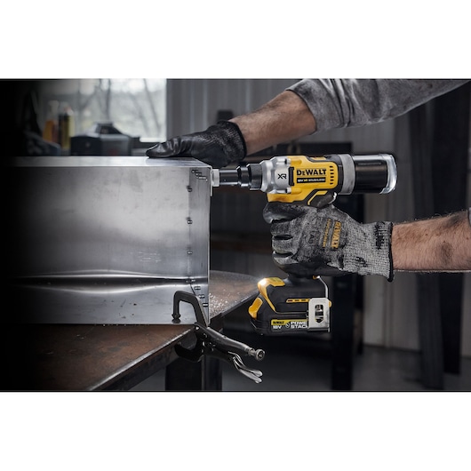 A person wearing work gloves is using a DEWALT DCF414NT 18V cordless rivet gun to fasten a metal sheet in an industrial workshop.