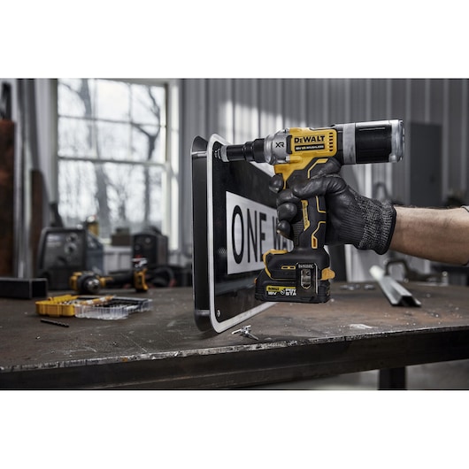 A person wearing a black glove uses a DEWALT DCF414NT cordless power tool on a metal sign in a workshop. The tool features yellow and black DEWALT branding and is positioned against the edge of the sign. Various tools and parts are visible on the workbench in the background.