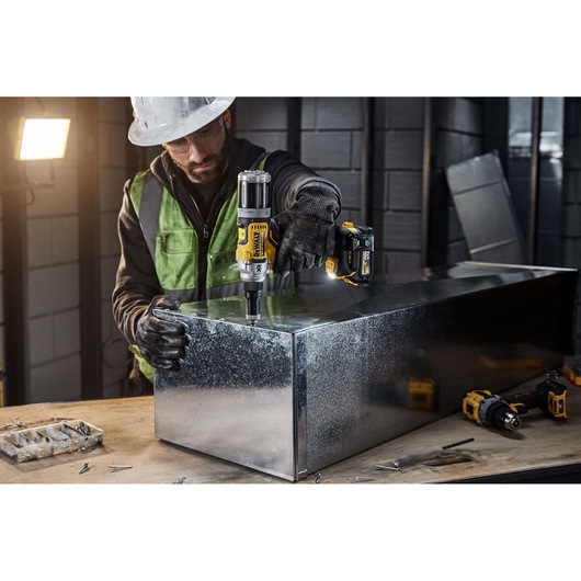 A person wearing protective gear uses a DEWALT DCF414NT cordless power tool to work on a large metal box in an industrial setting. Various tools and screws are visible on the worktable.
