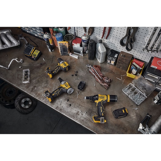 A workshop scene with DEWALT power tools, including a cordless screwdriver and drill, placed on a metal workbench surrounded by various accessories, screws, batteries, charger, and hand tools. The background includes shelves with containers and spare parts.