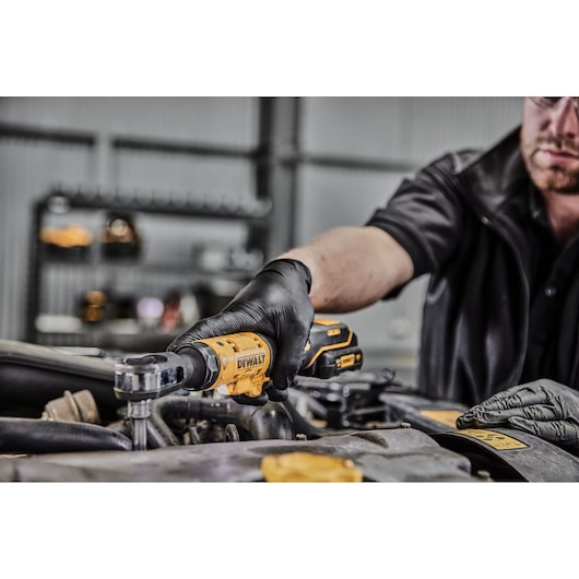 A person wearing black gloves uses a yellow DEWALT cordless ratchet tool to work on an engine in a workshop.