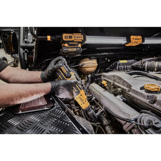 A person wearing black gloves uses a yellow DEWALT DCF503N cordless ratchet wrench to work on an engine inside a vehicle. Other DEWALT tools and equipment are visible mounted under the raised hood.