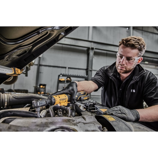 A person wearing black gloves uses a DEWALT DCF504L1G cordless ratchet tool to work on a car engine inside a garage.