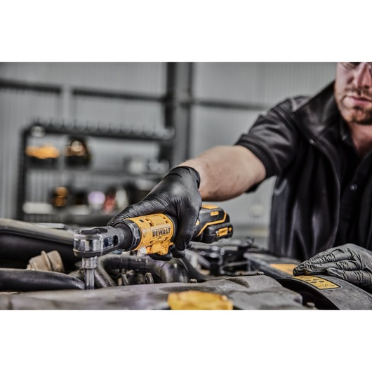 A person wearing black gloves uses a DEWALT DCF504N cordless ratchet tool to work on a car engine in a garage setting.