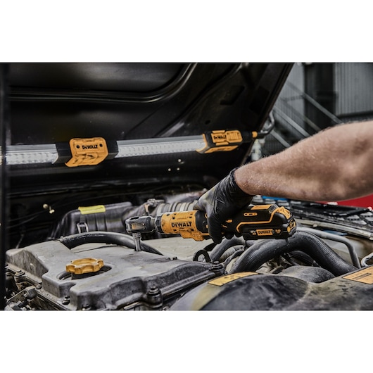 A person uses a DEWALT cordless ratchet tool (model DCF504N) to work on a car engine in a garage setting. An LED work light with DEWALT branding is attached to the car hood for illumination.