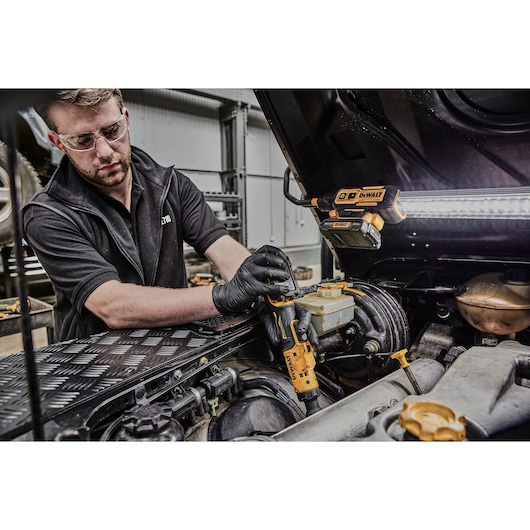A person wearing black gloves uses a DEWALT power tool, likely a cordless ratchet, to work on the engine of a vehicle in a workshop. A DEWALT work light is attached to the underside of the open car hood, illuminating the work area.