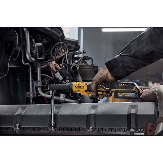 A person uses a DEWALT cordless ratchet tool to work on automotive engine components, showing the tool powered by an 18V POWERSTACK battery.
