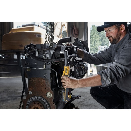 A person in work clothing uses a DEWALT cordless ratchet tool to tighten bolts on a large engine component inside a workshop. The person’s face is blurred for privacy.