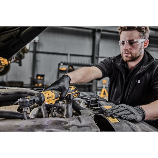 Person wearing black gloves using a DEWALT DCF512 cordless ratchet wrench to work on a car engine in a workshop.