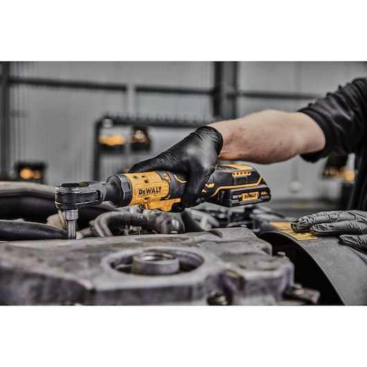 A person wearing black gloves uses a DEWALT cordless ratchet tool, model DCF512D1G, to work on a mechanical component, possibly in an automotive setting.