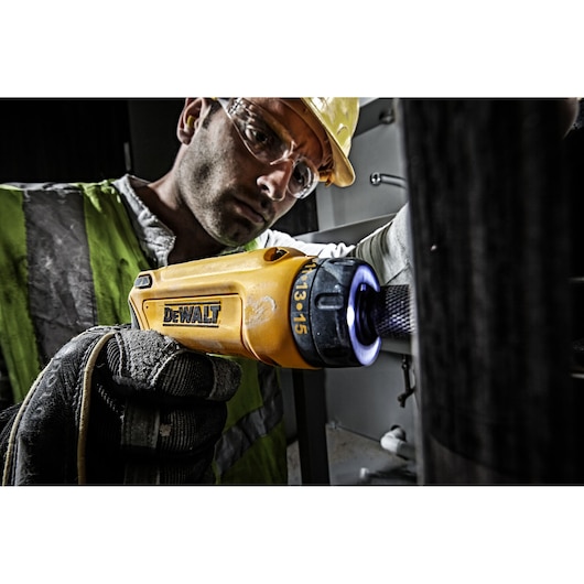 A person wearing a reflective safety vest and gloves uses a yellow DEWALT cordless screwdriver (model DCF680G2) for maintenance work indoors. The focus is on the tool and the user's gloved hand. The person's face is blurred for privacy.