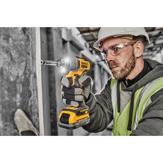 A person wearing work gloves, a safety helmet, and a high-visibility vest is using a DEWALT DCF840 cordless impact driver to drive a screw into a vertical surface on a construction site.