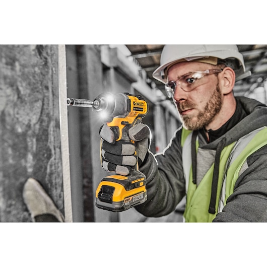 A person wearing safety gear uses a DEWALT DCF840N cordless impact driver to fasten a screw to a wall in a construction setting. The person’s face is blurred for privacy.