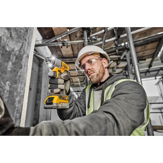A person wearing work gloves, a hard hat, and a high-visibility vest is using a DEWALT DCF840N cordless impact driver to drive a screw into a metal surface on a construction site. The tool is yellow and black, and the work environment contains scaffolding and metal structures.