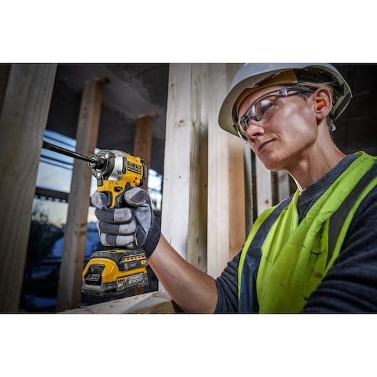 A person wearing a safety vest, gloves, and a hard hat is using a DEWALT DCF850E1T cordless impact driver at a construction site, holding the tool against a wooden frame.