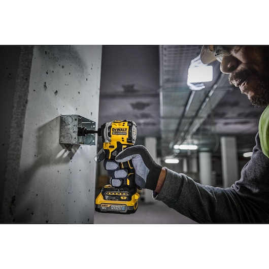 A person wearing safety gloves uses a DEWALT cordless impact driver (model DCF850) to fasten a screw into a concrete wall in an industrial or construction setting.