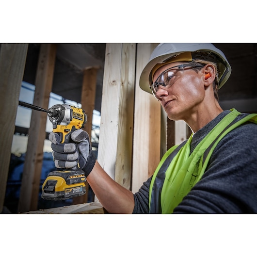Person wearing a hard hat and high-visibility vest using a DEWALT cordless power tool, specifically an impact driver, on a construction site.