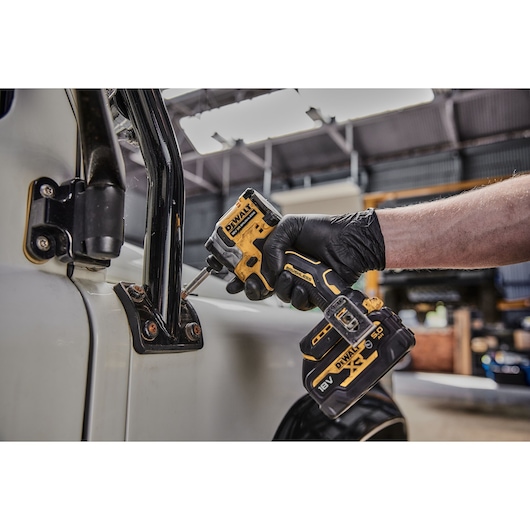 A person wearing a black glove uses a DEWALT cordless impact driver to fasten a screw on a metal hinge attached to a vehicle door in a workshop.