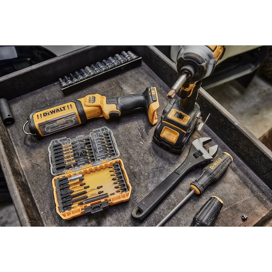 A selection of DEWALT tools, including a cordless impact driver, a screwdriver, an adjustable wrench, a set of drill bits in a yellow plastic case, and socket attachments, arranged on a workbench.