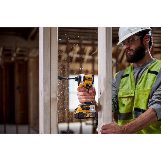 A person wearing a safety helmet and high-visibility vest uses a DEWALT DCF850N cordless drill to drill through a wooden frame in a construction setting.