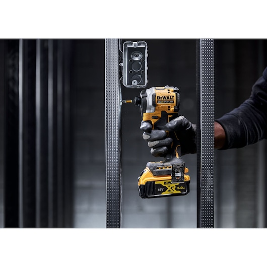 A person wearing black gloves uses a DEWALT cordless impact driver to screw into a metal frame near an electrical box.