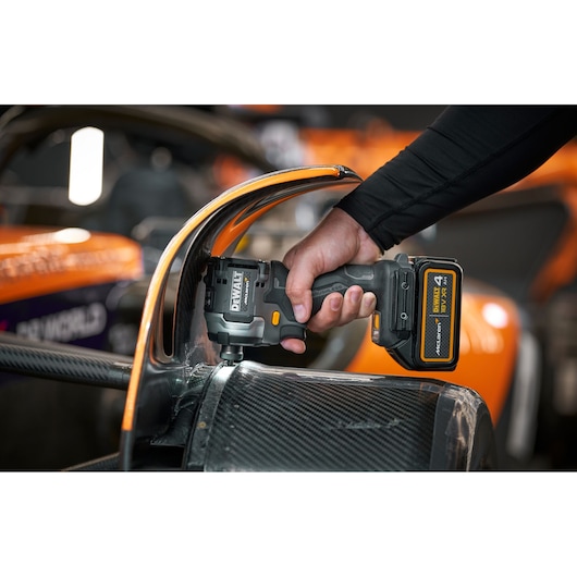A person uses a DEWALT cordless power tool, specifically a DCF85MM1T impact driver, to work on the body of a race car with orange and black details.
