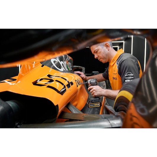 A person wearing an orange and black uniform is using a DEWALT power drill on the side of an orange race car with visible sponsorship logos. The person's face is blurred.