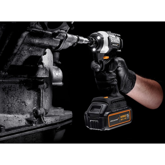 A gloved hand using a DEWALT McLaren 18V cordless impact driver with a 5Ah battery to work on a metal machine part. The tool is illuminated by built-in LED lights.
