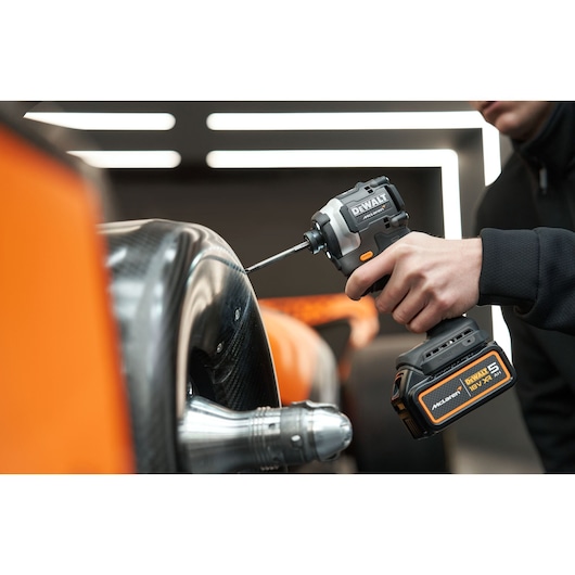 A person using a DEWALT cordless impact driver to fasten a screw on a carbon fiber automotive part. The tool features McLaren branding and an 18V XR 5.0Ah battery.