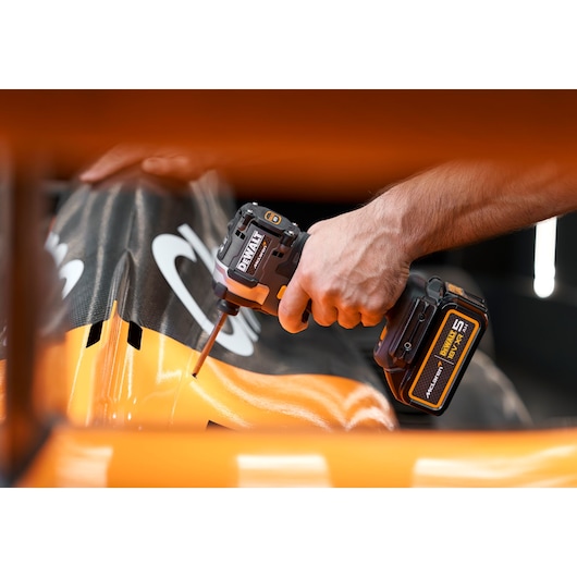 A person using a DEWALT cordless impact driver to work on an orange and black surface, possibly part of a vehicle or machinery.