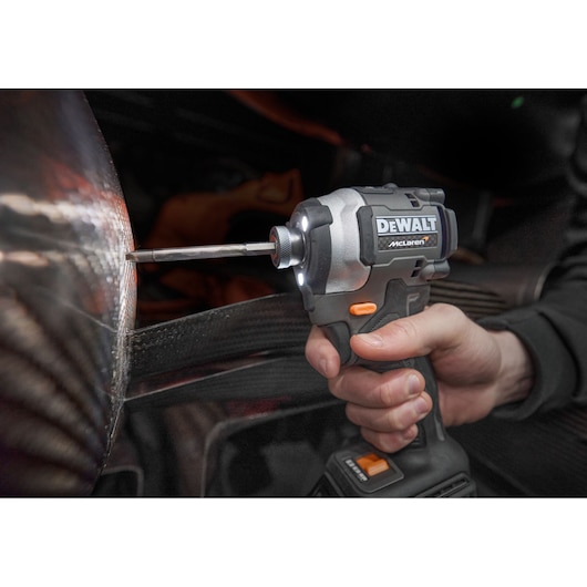 A person is using a DEWALT McLaren cordless impact driver to work on a metallic surface, with the tool's LED light illuminating the area.