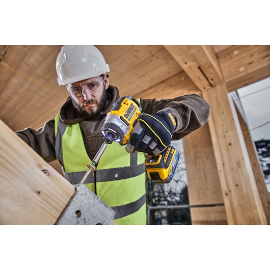 A person wearing a safety helmet, gloves, and a high-visibility vest is using a DEWALT DCF860E2T cordless impact driver to fasten a screw into a wooden beam at a construction site.