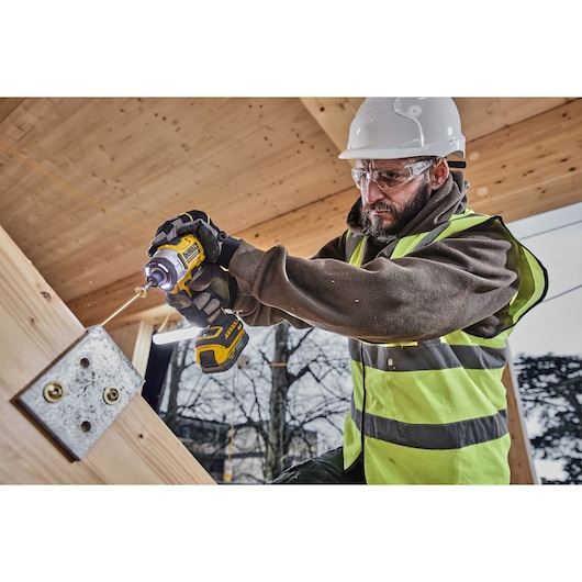 A construction worker wearing a safety vest and hard hat is using a DEWALT cordless impact driver to drill a screw into a metal bracket attached to a wooden beam. The tool is clearly visible and in use in an outdoor worksite setting.