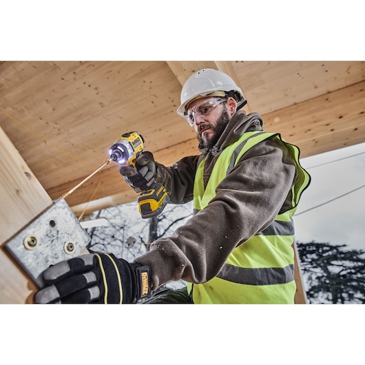 A person wearing a safety helmet, gloves, and a high-visibility vest is using a DEWALT cordless impact driver (DCF860E2T) to drill into a metal plate attached to a wooden structure at a construction site.