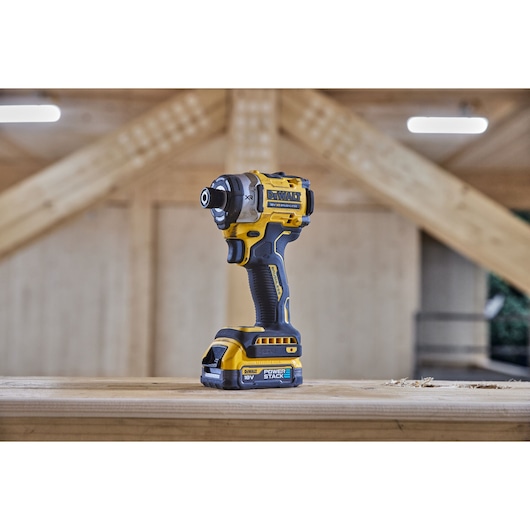 A DEWALT 18V cordless impact driver tool standing upright on a wooden surface in a workshop setting, with exposed wooden beams and blurred background.