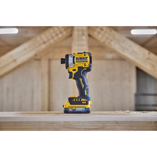 A DEWALT DCF860E2T 18V cordless impact driver with Powerstack battery is standing upright on a wooden surface inside a workshop with wooden beams in the background.