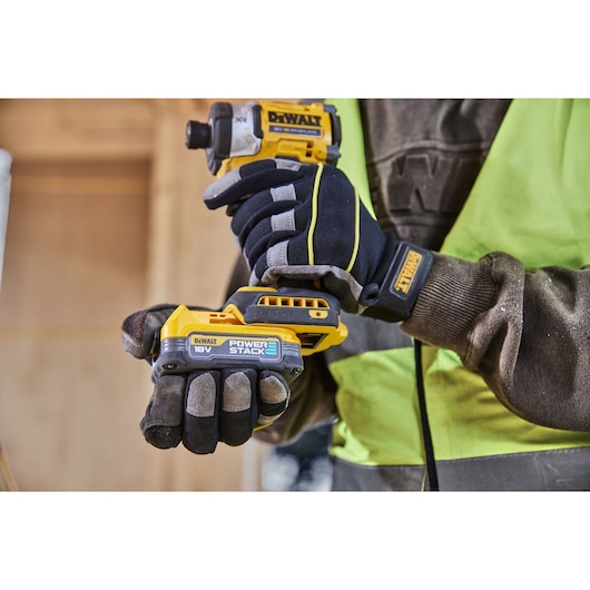 A close-up of a person wearing work gloves and a high-visibility vest, holding a DEWALT 18V Power Stack cordless impact driver in a construction or workshop setting.