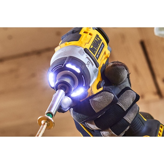 Close-up view of a DEWALT DCF860E2T cordless impact driver being used, with LED lights illuminating the working area. The tool is held by a person wearing protective gloves.