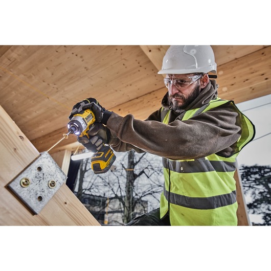 A person wearing a high-visibility vest and a hard hat is using a DEWALT cordless impact driver (model DCF860NT) to fasten a screw into a metal bracket attached to a wooden beam at a construction site.