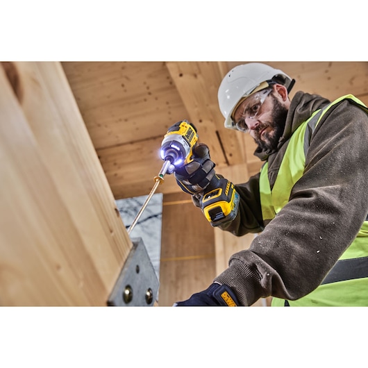 A person wearing a safety helmet and high-visibility vest is using a DEWALT cordless impact driver (model DCF860NT) to fasten a screw into a wooden structure at a construction site.