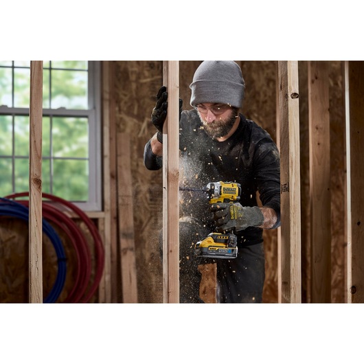 A person wearing gloves and a beanie is using a DEWALT power tool to drill or drive screws into a wooden frame, with sawdust flying in a construction setting. The face of the person is blurred.