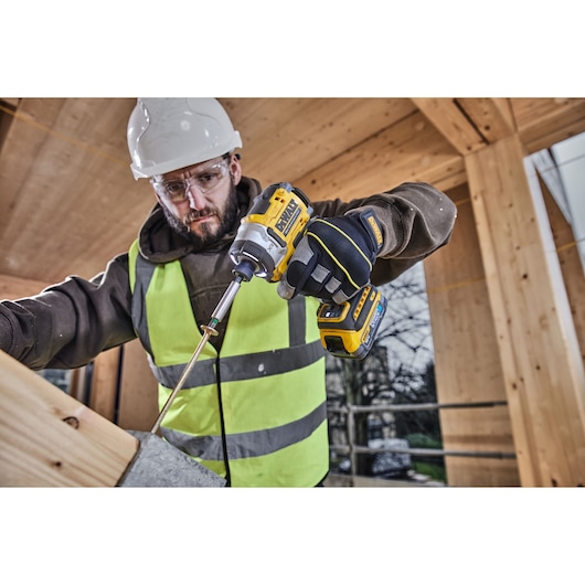 A person wearing a high-visibility vest, gloves, and a hard hat is using a DEWALT cordless impact driver (model DCF860N) to drive a screw into a wooden structure at a construction site. The background shows wooden beams and an outdoor environment.