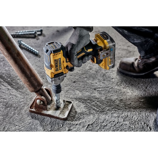 A person wearing work gloves and boots using a DEWALT cordless impact wrench to tighten a bolt on a rusty metal bracket attached to a concrete surface.