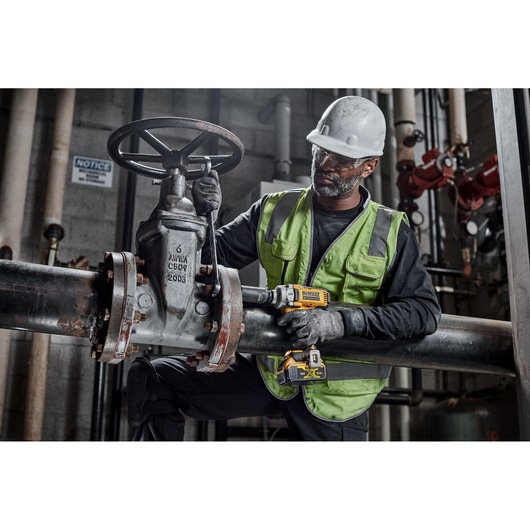 A worker wearing a safety helmet and reflective vest is using a DEWALT DCF891NT power tool on a large industrial pipe valve in a factory or plant setting. The worker's face is blurred for privacy.